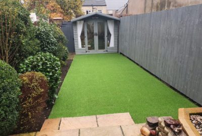 Photo of fake grass installation in Barnsley, with shrubs to the left, grey painted palisade fencing, and a grey painted summerhouse to the back