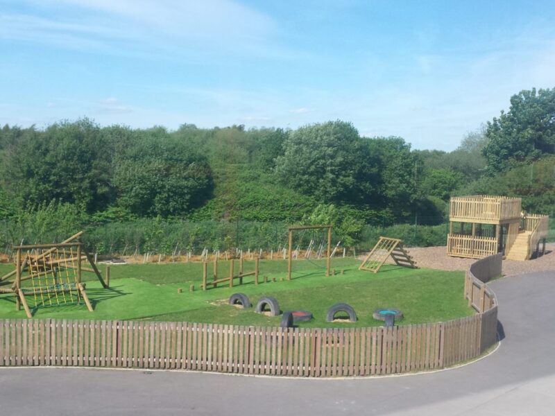 Photo looking out at school wooden playground equipment, featuring a timber trim trail and tangle climber installed on artificial turf safety surfacing, and a bespoke lookout tower over looking a river on play bark