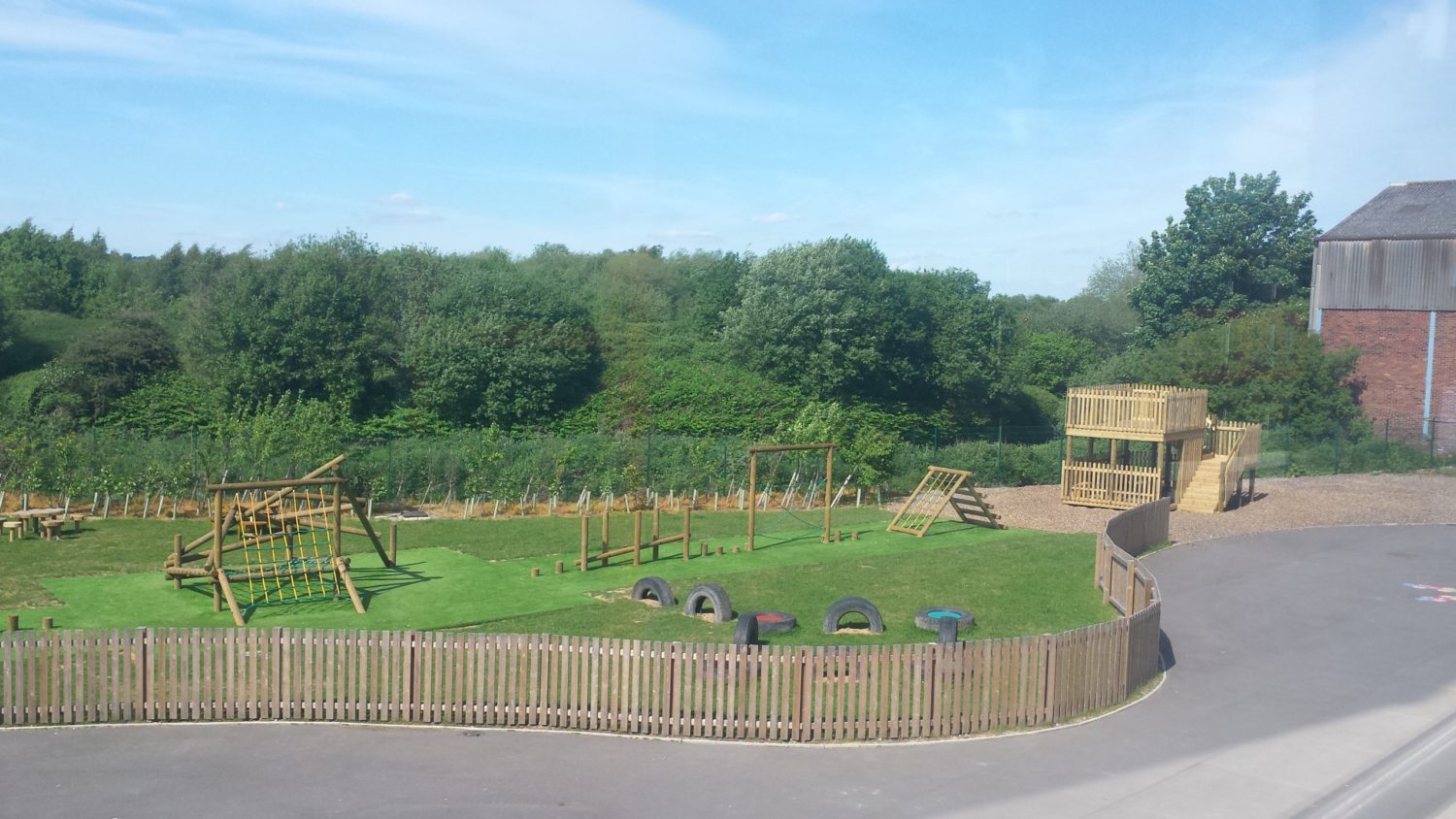 Photo looking out at school wooden playground equipment, featuring a timber trim trail and tangle climber installed on artificial turf safety surfacing, and a bespoke lookout tower over looking a river on play bark