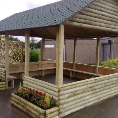 Brayton Log Cabin timber outdoor shelter for schools with all-round seating, waterproof roof, and built-in planters