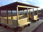 The Meadow Outdoor Classroom – timber shelter with pergola-style roof beams, boarded and felt roof, hit-and-miss fencing, and seating inside and outside.