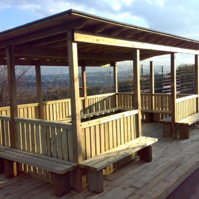 The Meadow Outdoor Classroom – timber shelter with pergola-style roof beams, boarded and felt roof, hit-and-miss fencing, and seating inside and outside.